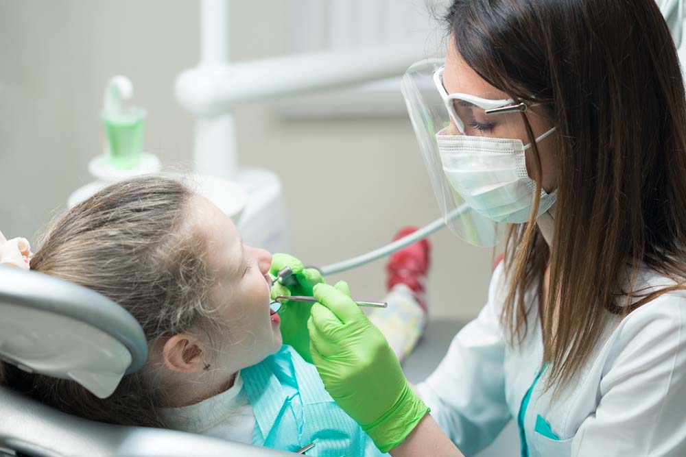 Pediatric dentist wearing a mask and face shield examining a young child’s teeth in a dental chair, using dental tools while the child looks up calmly.